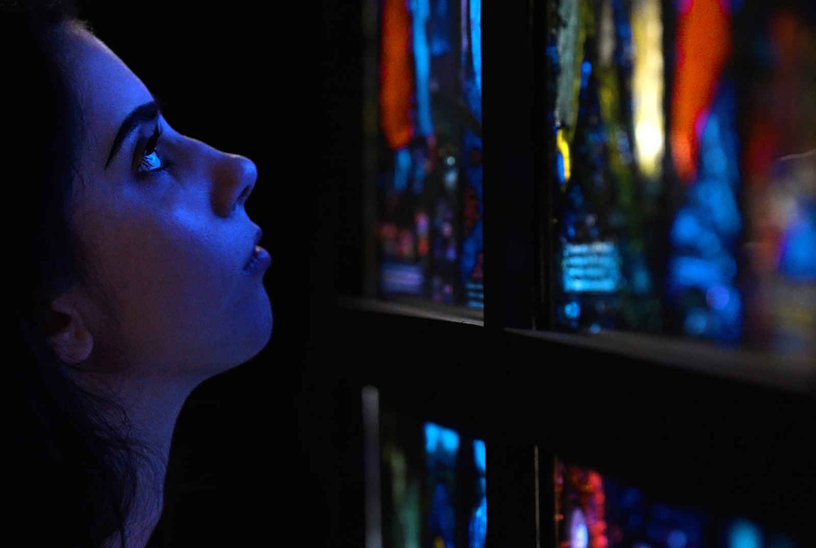 A person with face in profile in a dark gallery looking closely at a stained-glass window