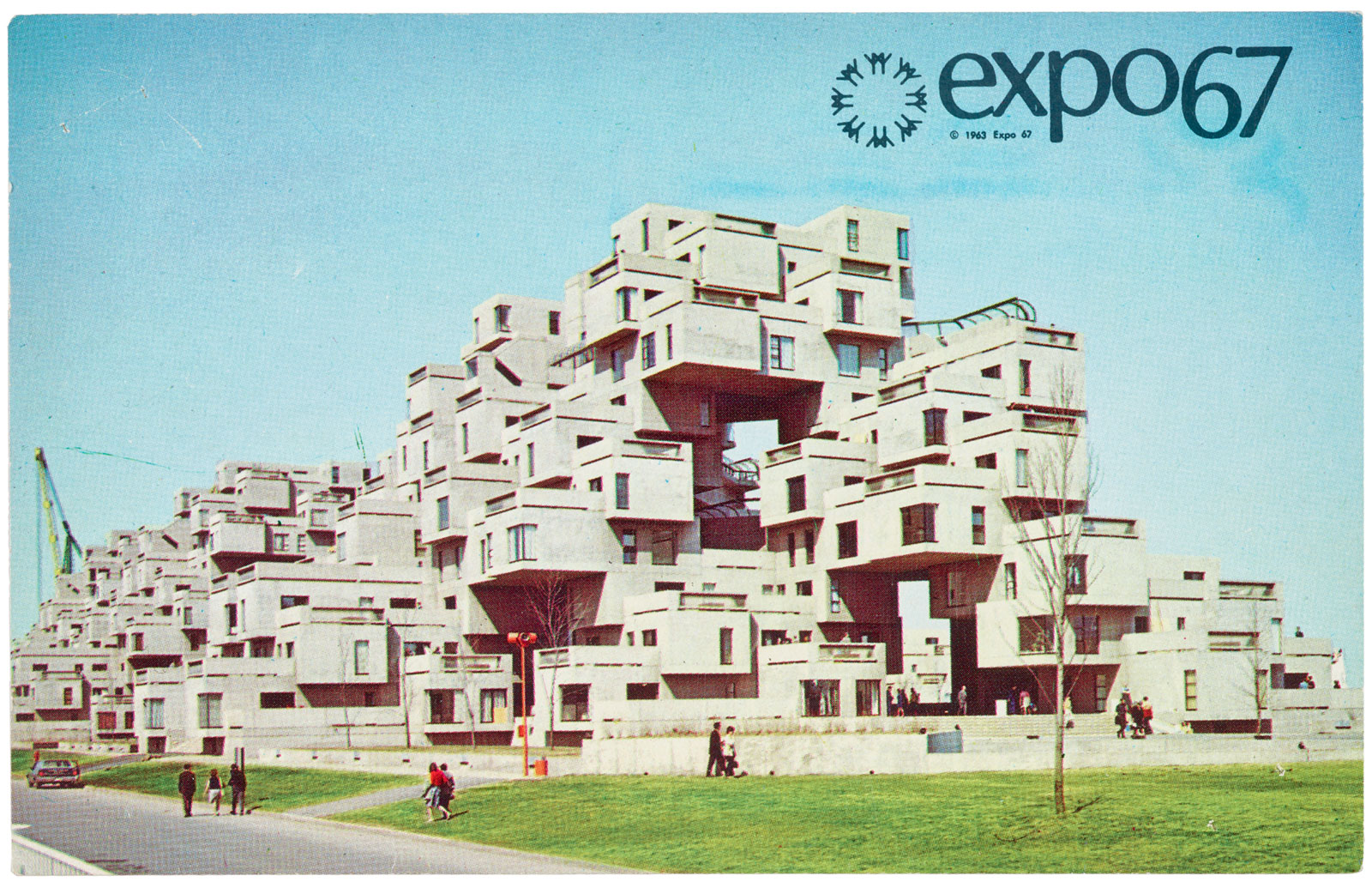 A postcard with “expo67” printed in the top right corner, depicting a large modular concrete housing complex composed of stacked rectangular units with windows and set on a grassy area under a clear blue sky, with people walking and standing nearby