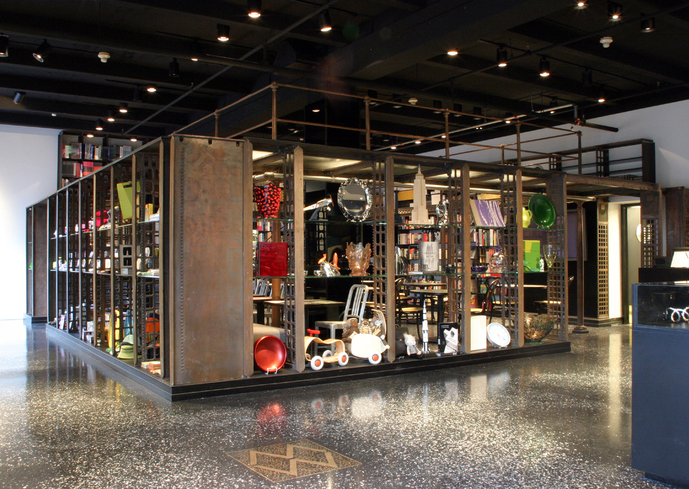 An open metal shelving unit filled with various objects including books, toys, housewares, and decorative items, located in a room with a black ceiling and speckled terrazzo floor