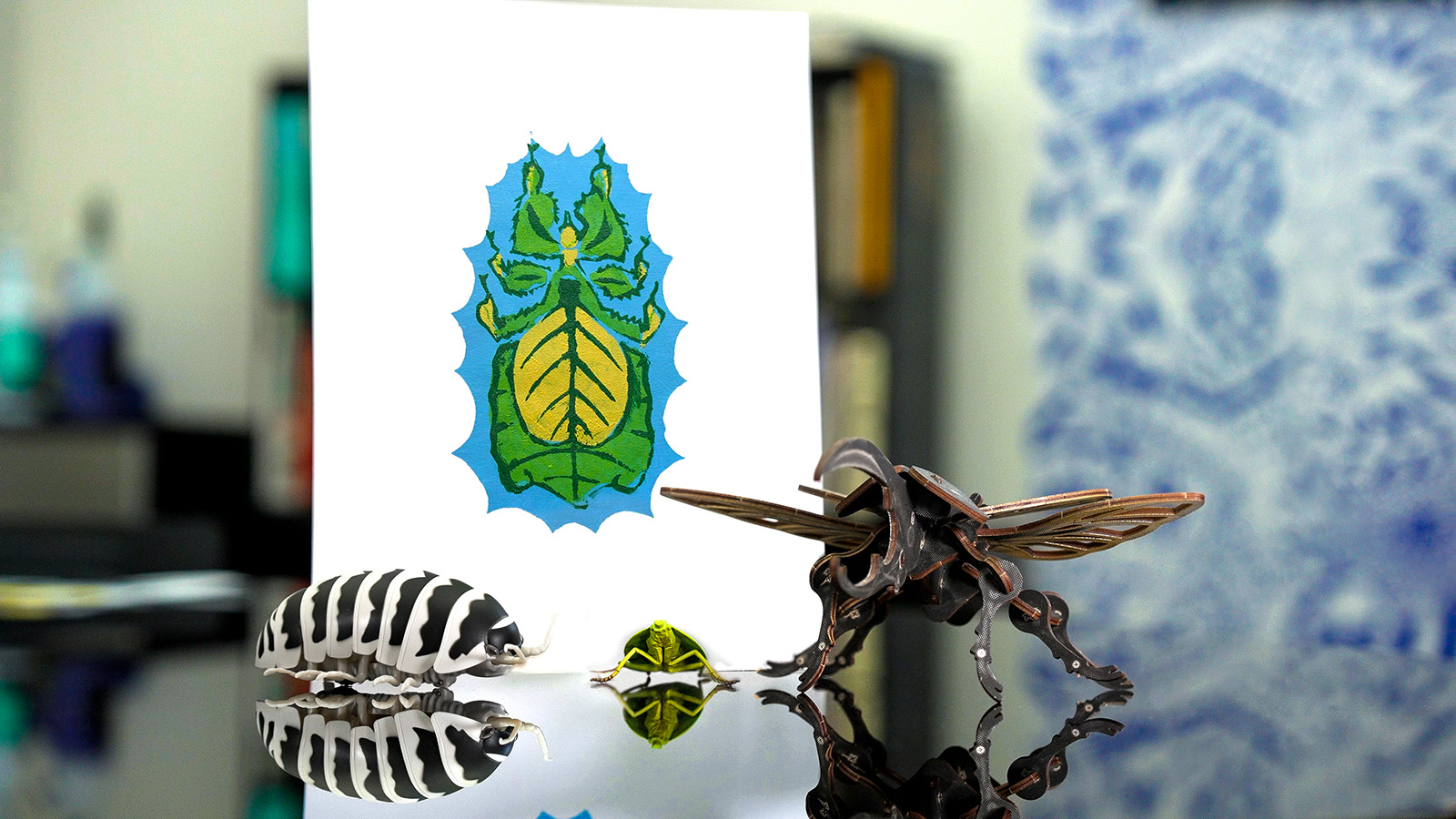A finished pochoir-style print of a green and yellow leaf insect with a blue scalloped border is displayed upright, surrounded by three insect figurines reflected on a glossy black surface