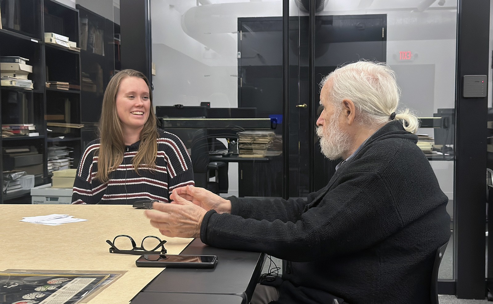 Two people at a table in an office setting are interacting and smiling. There are documents, a pair of glasses, and a smartphone on the table. Shelves and a glass wall are in the background.