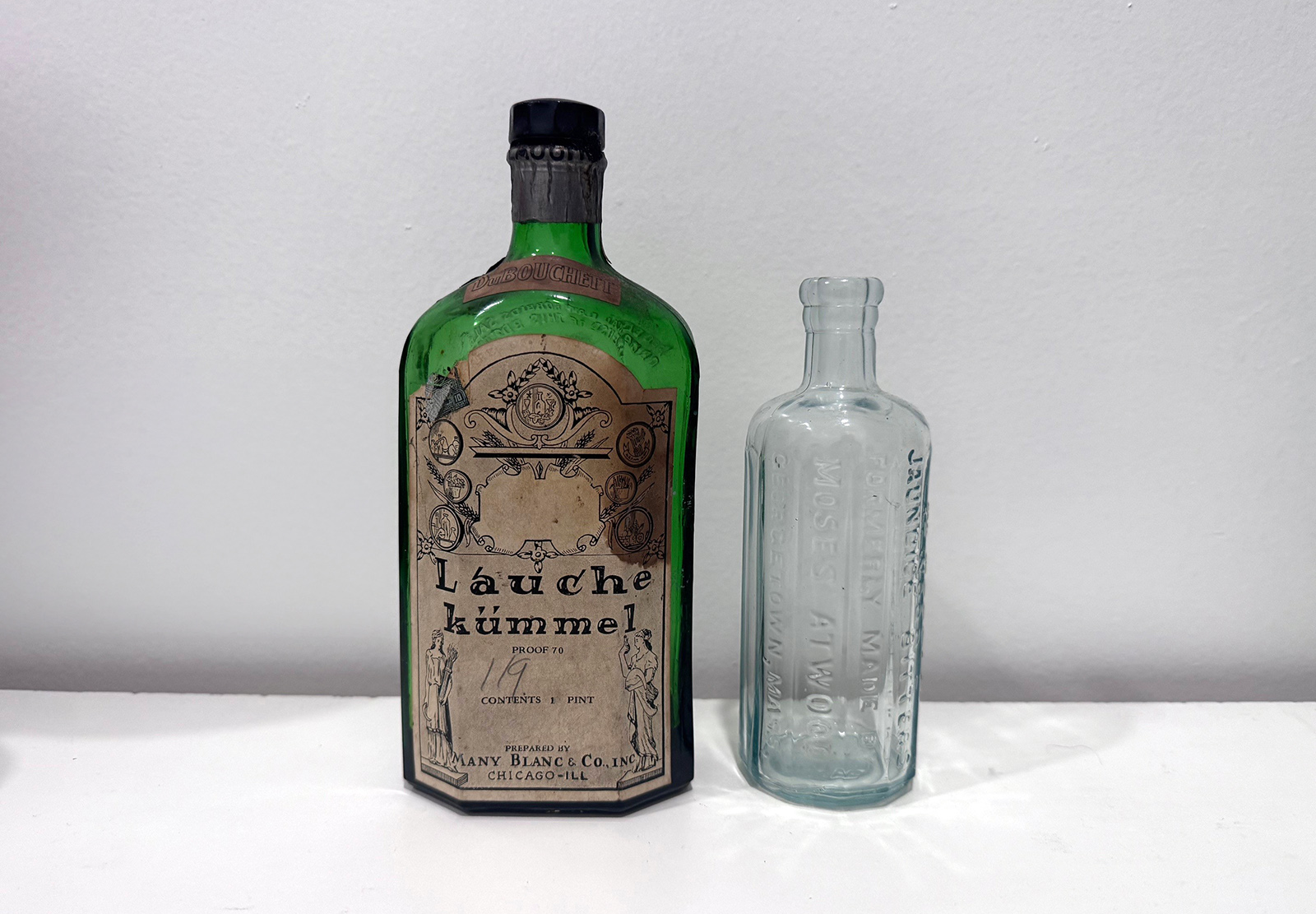 Two glass bottles, one green with a vintage label on it, and the other clear, stand on a table against a wall.