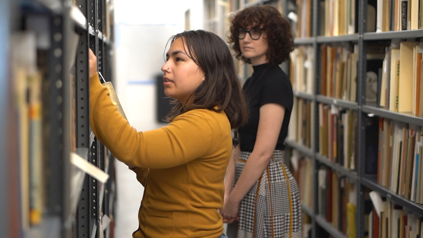 Two women stand between tall library shelves; one reaches for a book while the other observes from behind.