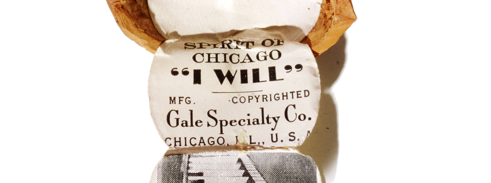 A partial, close-up view of a walnut shell containing a folded paper booklet printed with the words "Spirit of Chicago / 'I WILL' / MFG COPYRIGHTED / Gale Specialty Co. / Chicago, IL, USA"