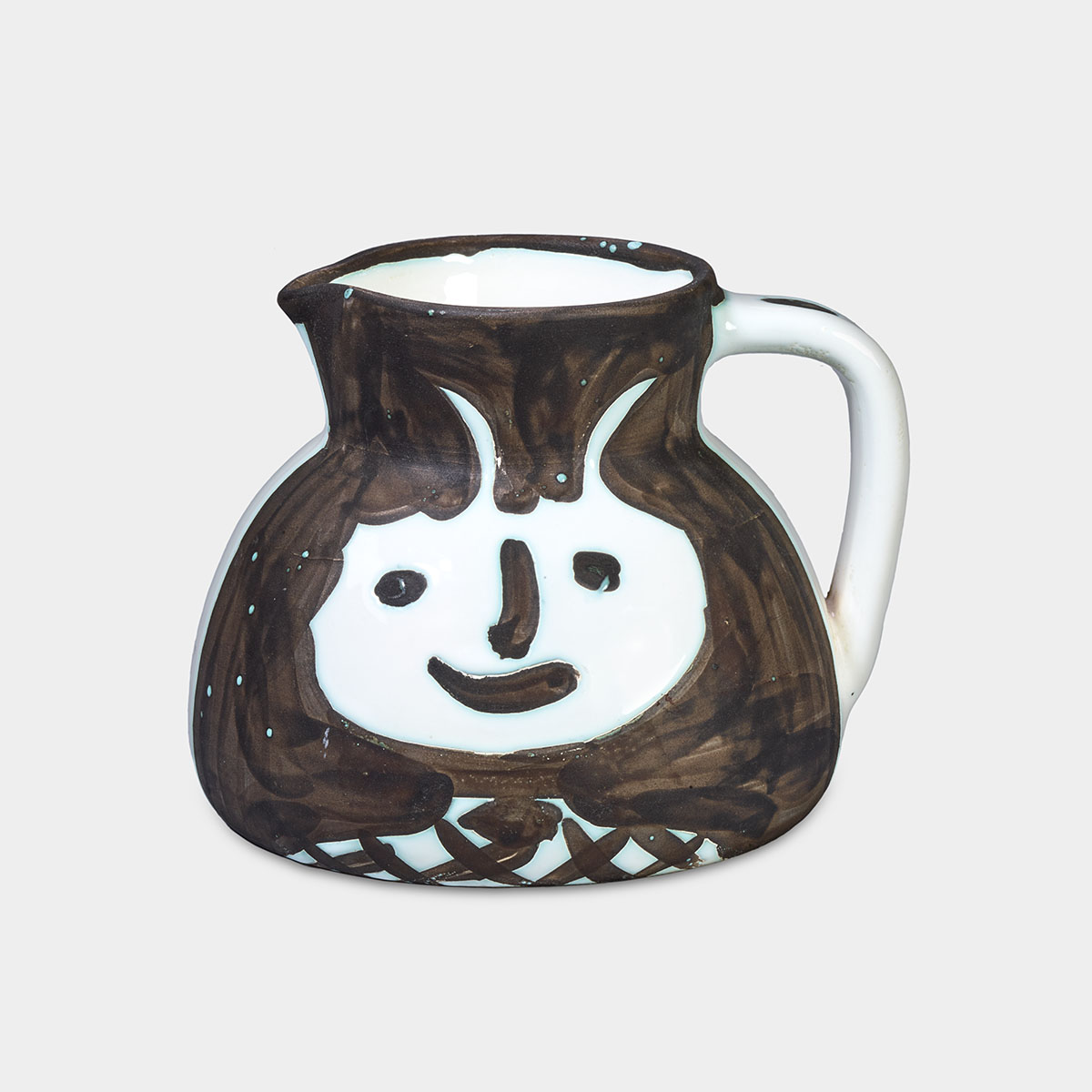 A smiling face appears on the surface of this ceramic pitcher. The side facing us is painted dark brown, while the area around the face is left unpainted, revealing the stark white color of the ceramic. The face is large and rounded, featuring wide, oval eyes, a dark brown line for a nose, and a curved mouth. Two horns protrude from the top of the head. Below the face, the neck and shoulders are white and centered at the base of the pitcher, adorned with thick, crossed dark brown lines. To our right, the handle of the pitcher has spots of dark brown paint along its length but remains mostly white. The mouth and rim at the top of the pitcher are painted dark brown, while the interior retains the same white color as the ceramic.