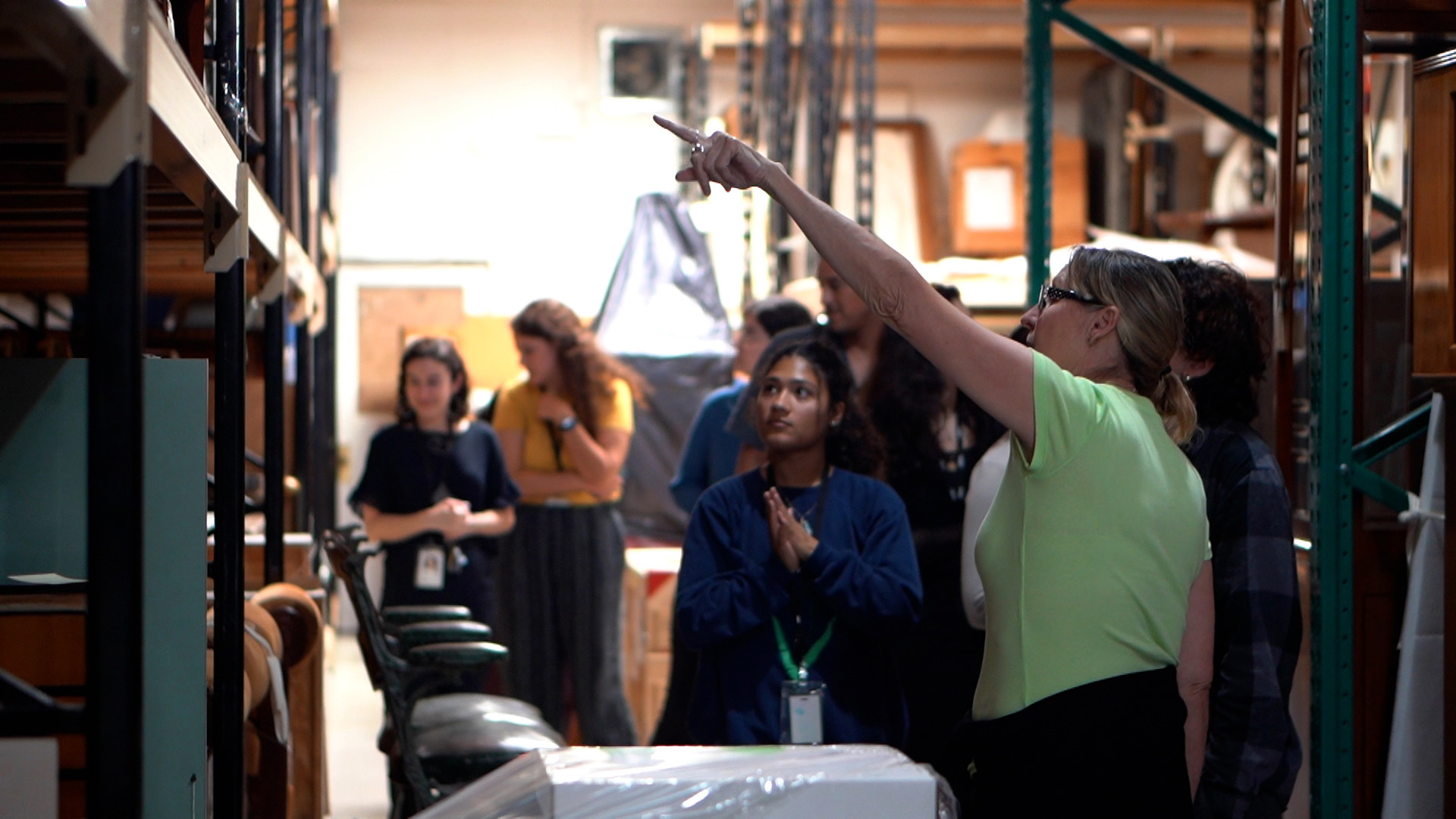 A woman gestures and points while leading a group through museum storage shelves lined with furniture and objects