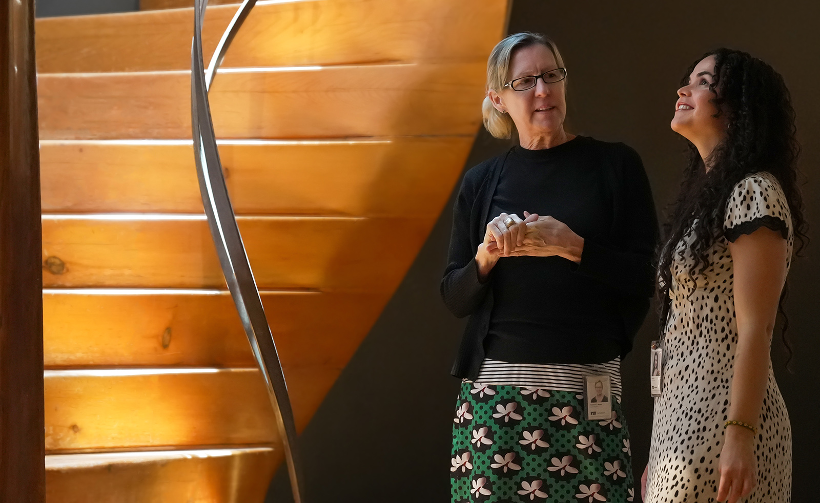 Two women stand talking near a large wooden spiral staircase, warmly lit from above