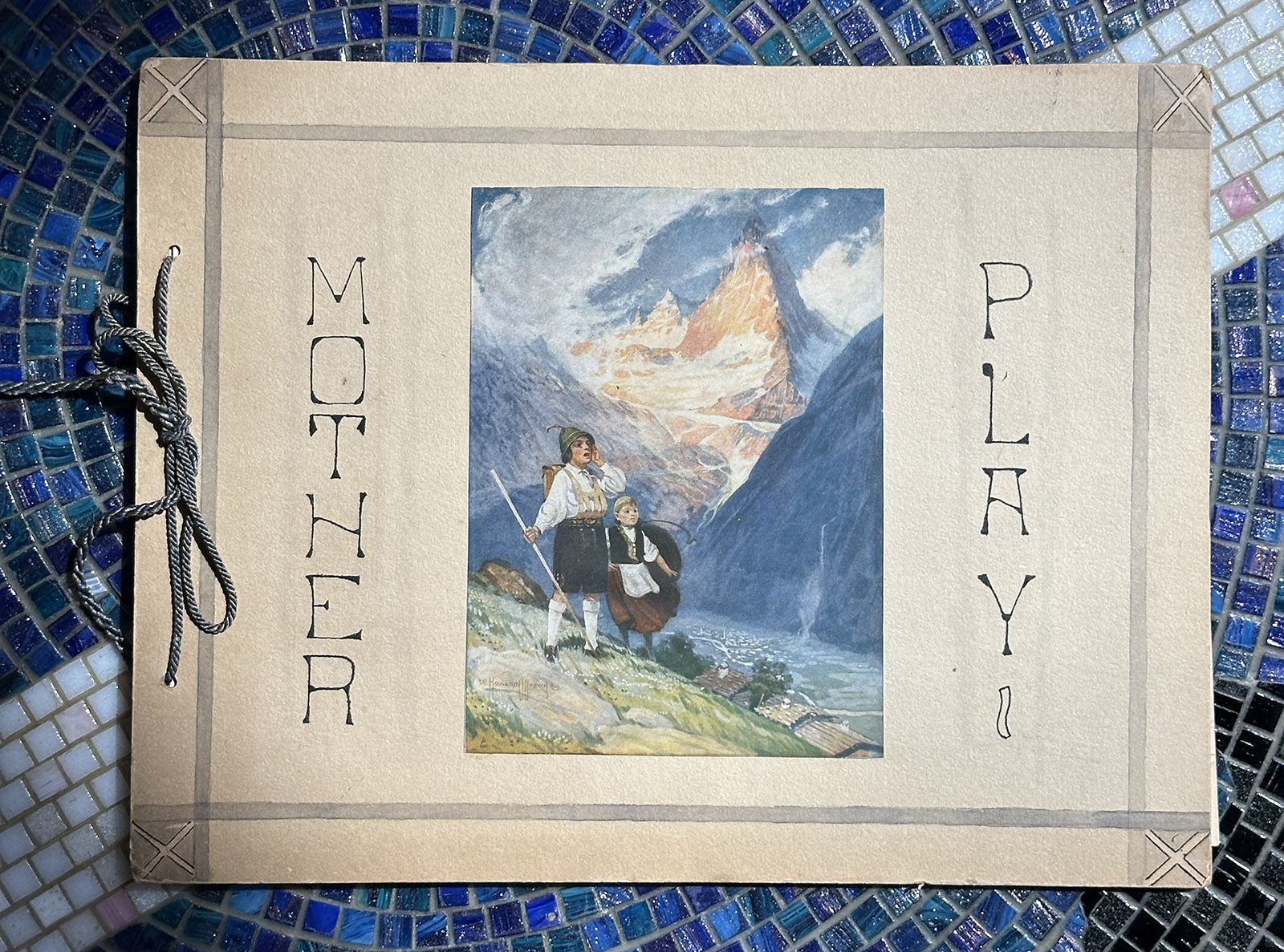 Cover of a booklet titled "Mother Play" with an alpine scene of a woman and child at center, bound with a cord and set against a blue mosaic background
